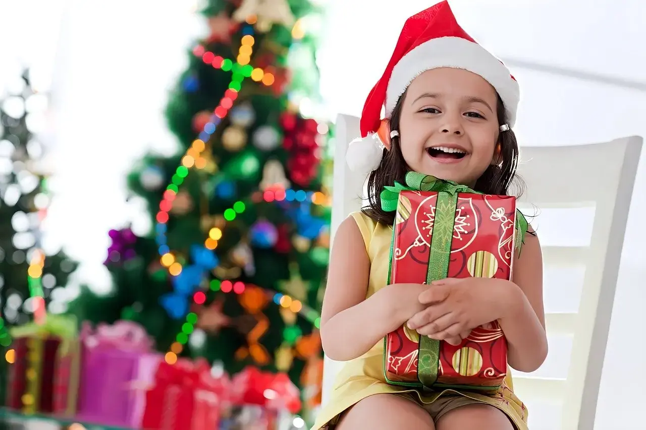  A young girl wearing a Santa hat joyfully holds a colorful gift in her hands, embodying the spirit of the holiday season
